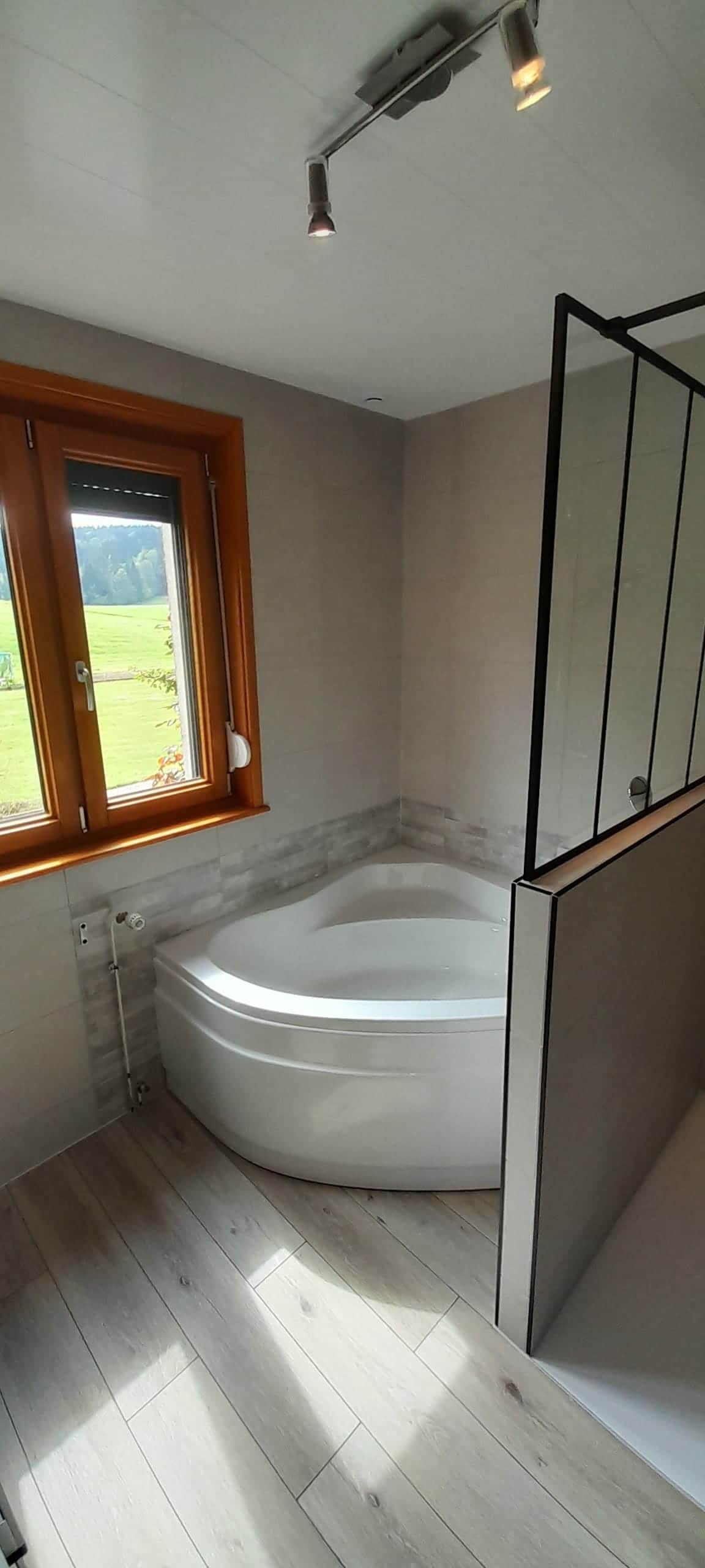 Salle de bain avec baignoire d'angle, paroi verrière noire et sol imitation parquet clair, par AML Plomberie