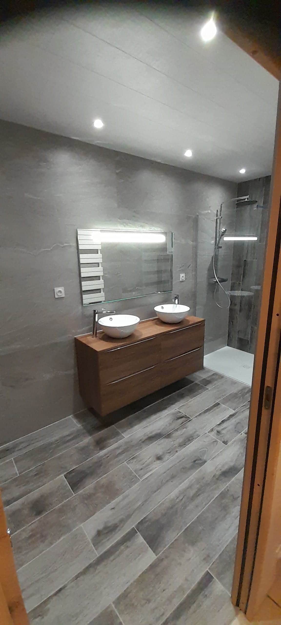 Salle de bain grise avec deux vasques à poser sur meuble bois suspendu, miroir lumineux et douche vitrée